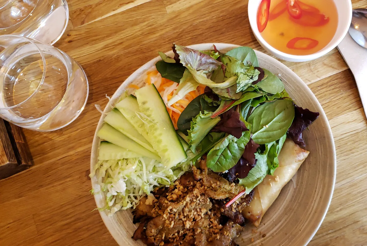 Schale mit gegrilltem Fleisch, Reisfadennudeln, Salat, Gurke und Frühlingsrolle, daneben eine Schale Dipsauce