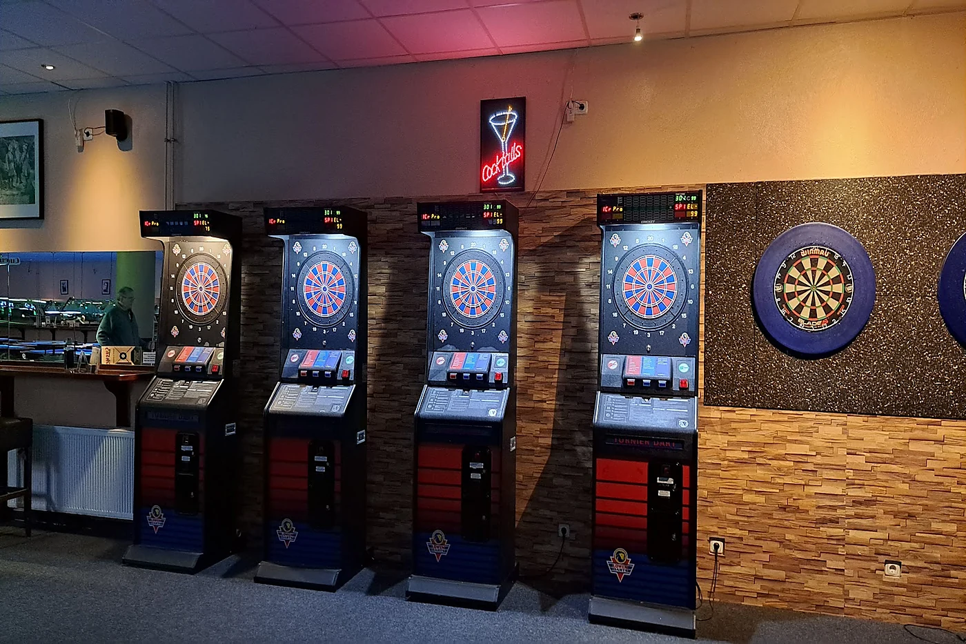 Reihe elektronischer Dartautomaten vor einer Steinwand, daneben eine klassische Dartscheibe und ein Neonschild