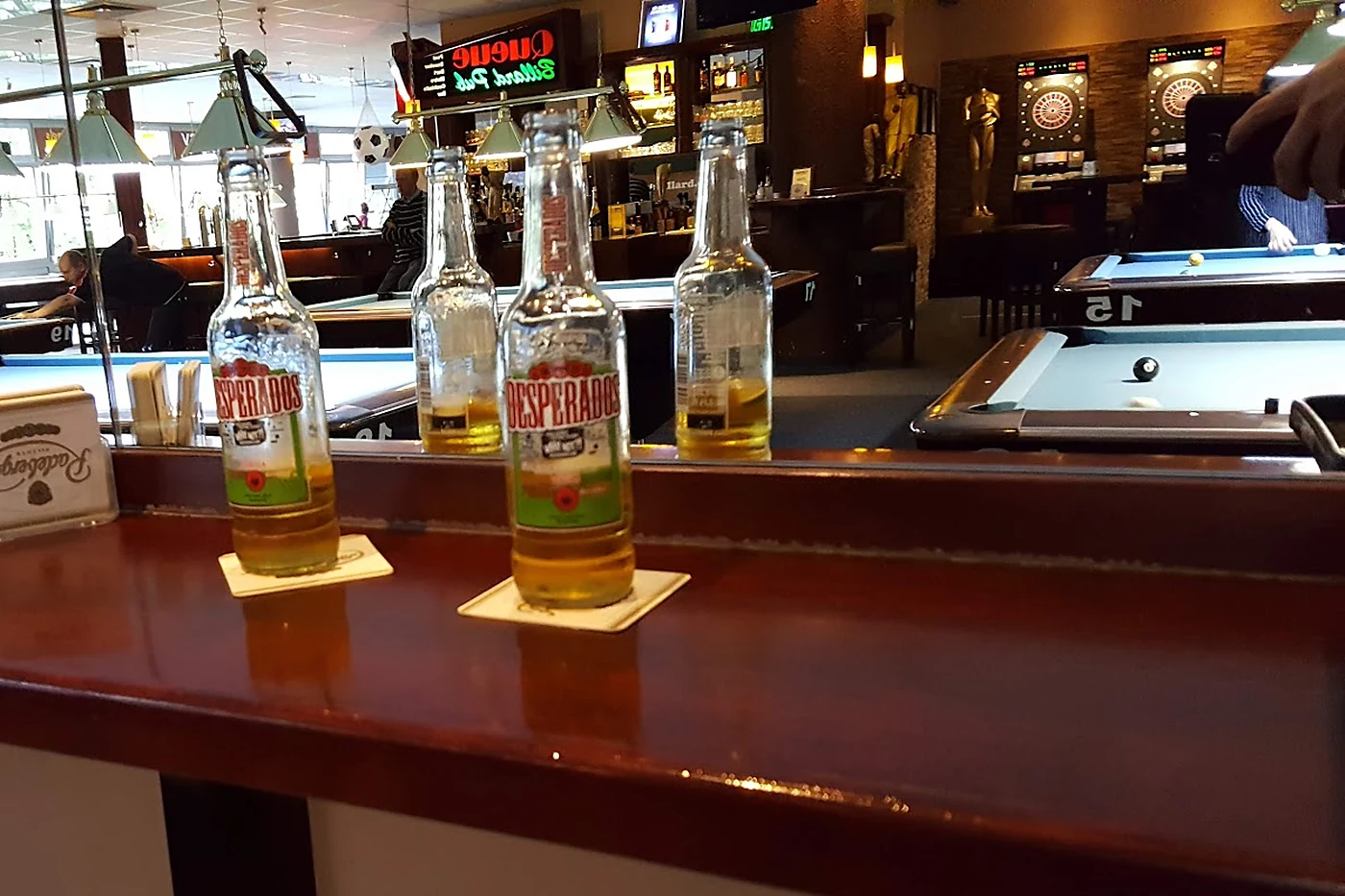 Bierflaschen auf Deckeln auf dem dunklen Holztresen, im Hintergrund Billardtische und Dartautomaten