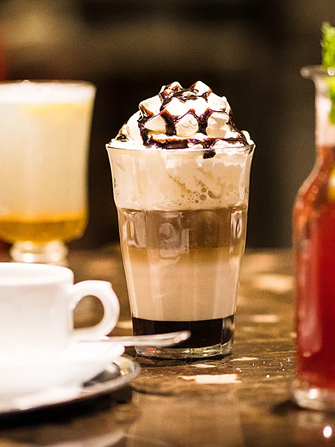 Eisschokolade mit Sahne und Schokoladensauce im hohen Glas, daneben eine Tasse Kaffee und ein rotes Getränk auf dem Tres