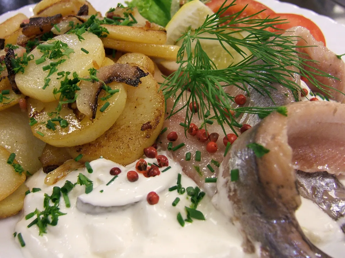 Matjesfilets mit Sahnesauce, Dill, rosa Pfeffer und Bratkartoffeln auf einem weißen Teller
