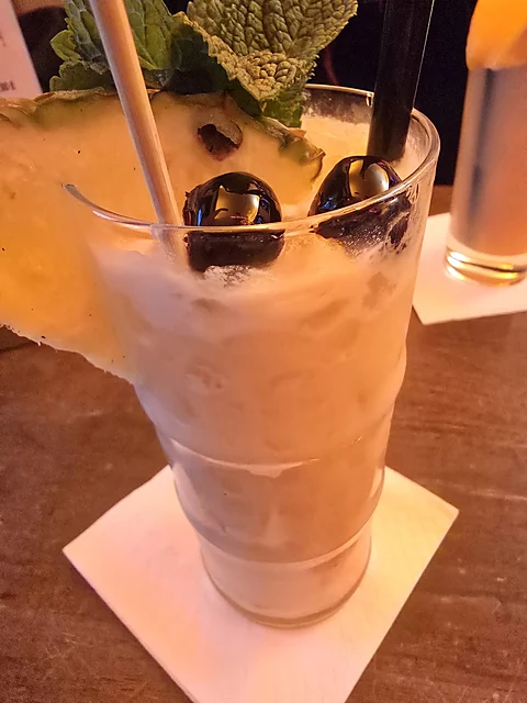 Cremiger Cocktail im hohen Glas mit Ananasstück, Minze und Kirschen auf dem Holztresen