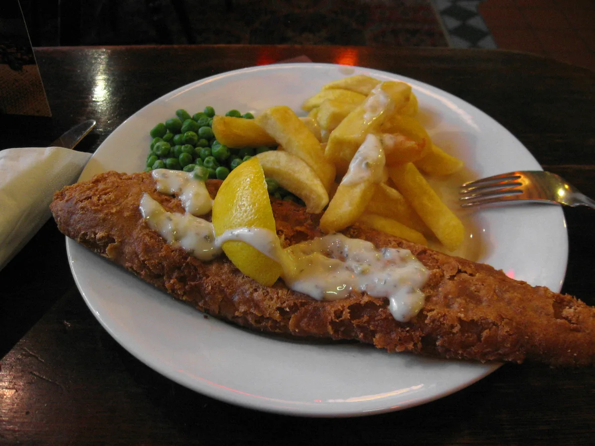 Paniertes Fischfilet mit Pommes frites, Erbsen, Zitrone und heller Sauce auf einem weißen Teller