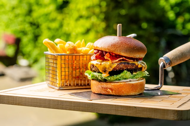Cheeseburger mit Bacon und Pommes im Drahtkörbchen auf einem Holzbrett im Biergarten
