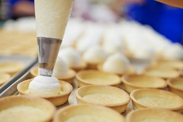 Ein Spritzbeutel füllt Creme in kleine Tartelette-Böden auf einem Backblech