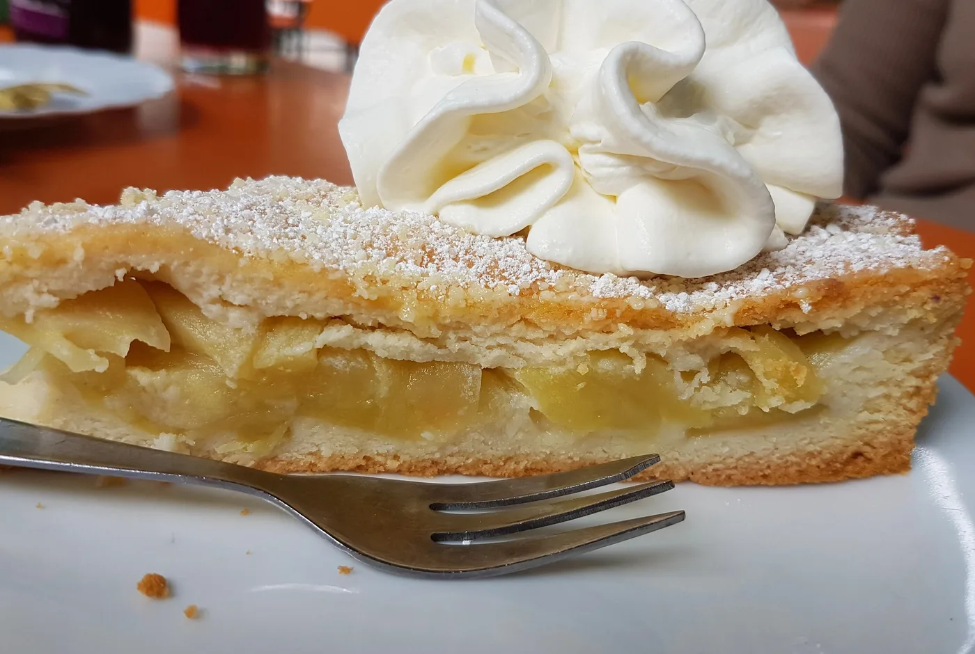Stück Apfelkuchen mit Puderzucker und einer großen Portion Schlagsahne auf einem weißen Teller