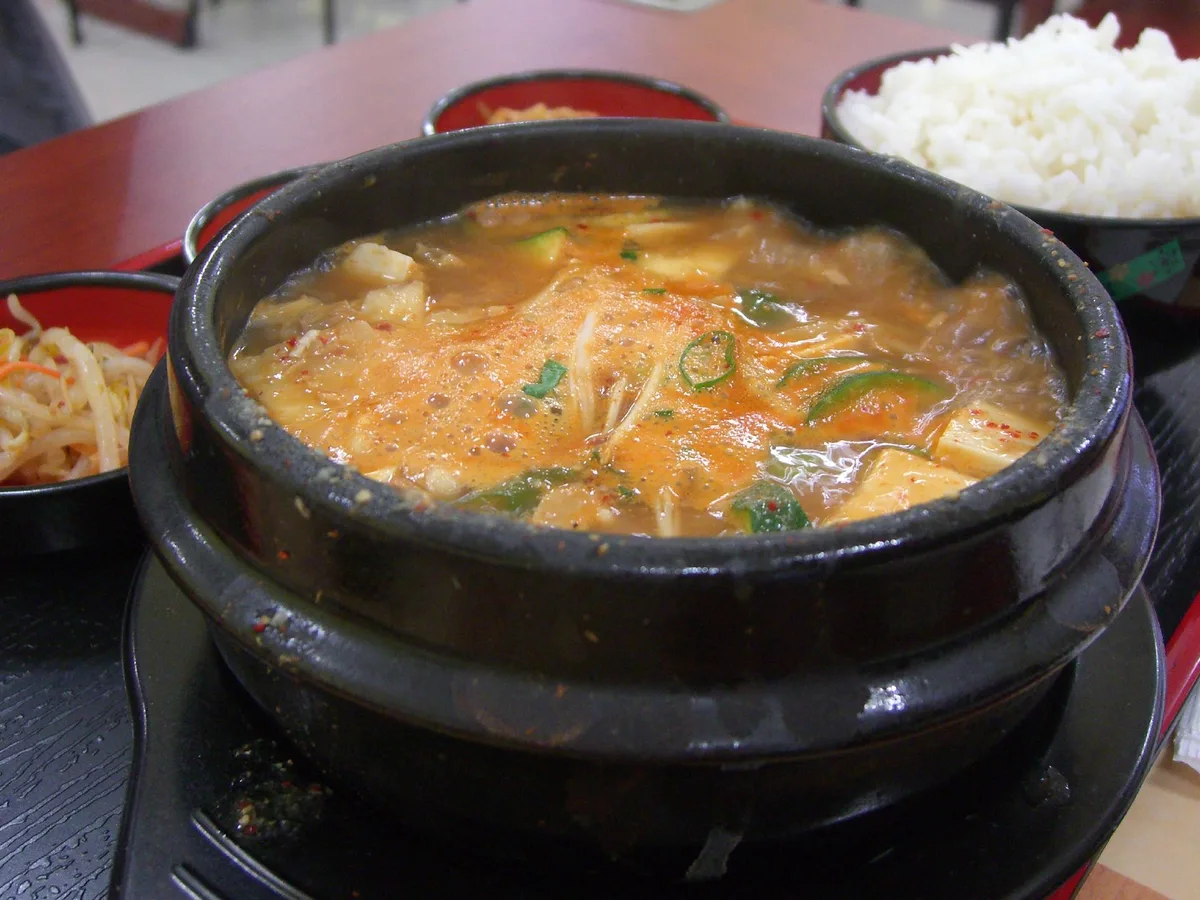 Koreanischer Eintopf im Tontopf mit Tofu und Gemüse, daneben Reis und Beilagen