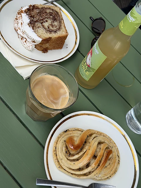 Tisch von oben: Kanelbulle auf goldrandigem Teller, Kaffee im Glas, ein Stück Streuselkuchen und eine Apfelschorle
