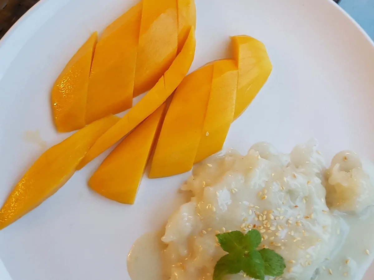 Aufgeschnittene Mango neben Klebreis mit Kokossauce, Sesam und Minze auf einem weißen Teller