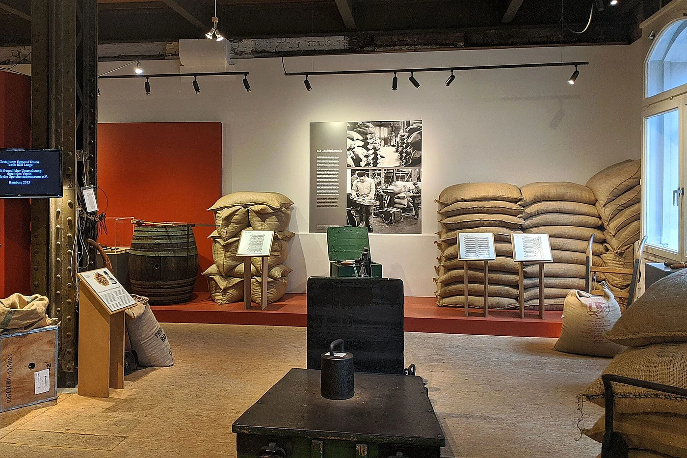 Ausstellungsraum mit gestapelten Kaffeesäcken, einem Holzfass, einer Sackkarre und historischen Fotos zwischen genietete