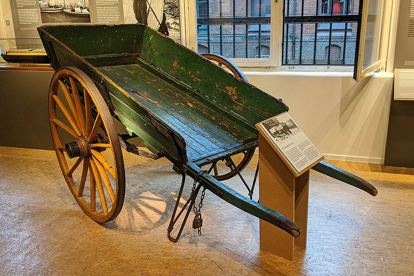 Grüne Holzkarre mit zwei großen Speichenrädern und langen Griffen auf dem Museumsboden, daneben eine Erklärtafel