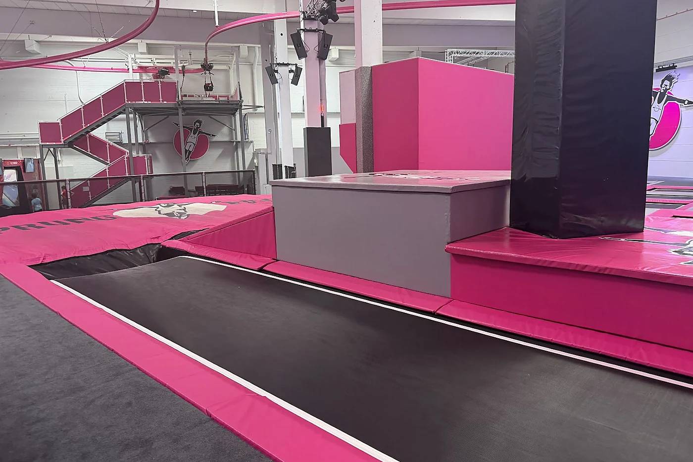Breite Trampolinbahn mit pinkfarbener Polsterung, dahinter Podeste, Treppen und eine schwarz gepolsterte Säule