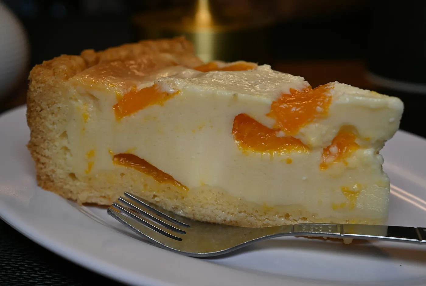 Stück Käsekuchen mit Pfirsichstücken auf einem weißen Teller, daneben eine Gabel
