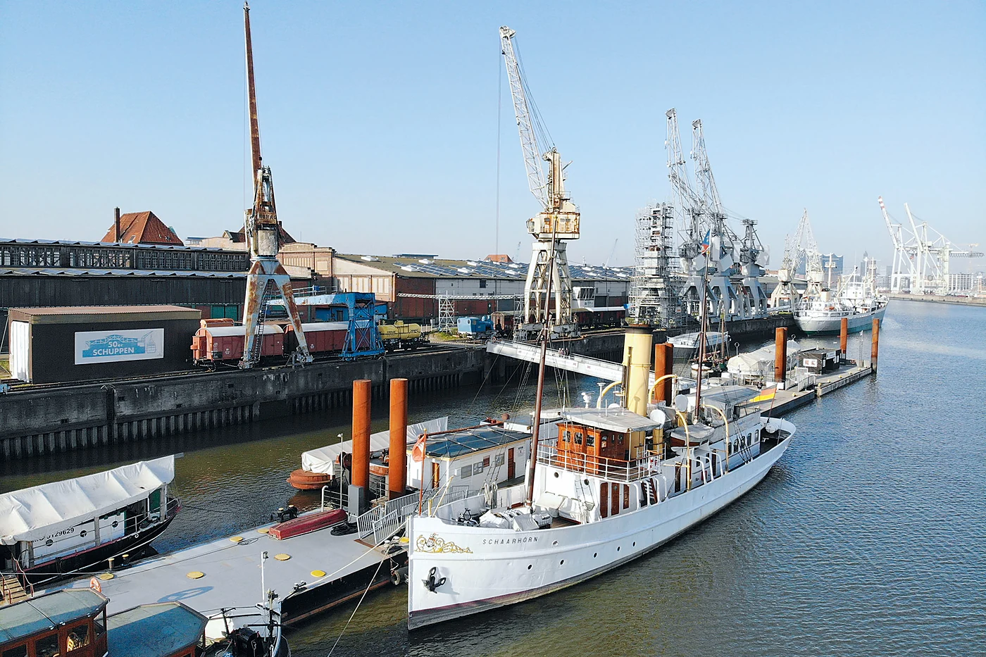 Der weiße Dampfer Schaarhörn und weitere historische Schiffe am Ponton des Bremer Kais, dahinter Hafenkrane und die 50er