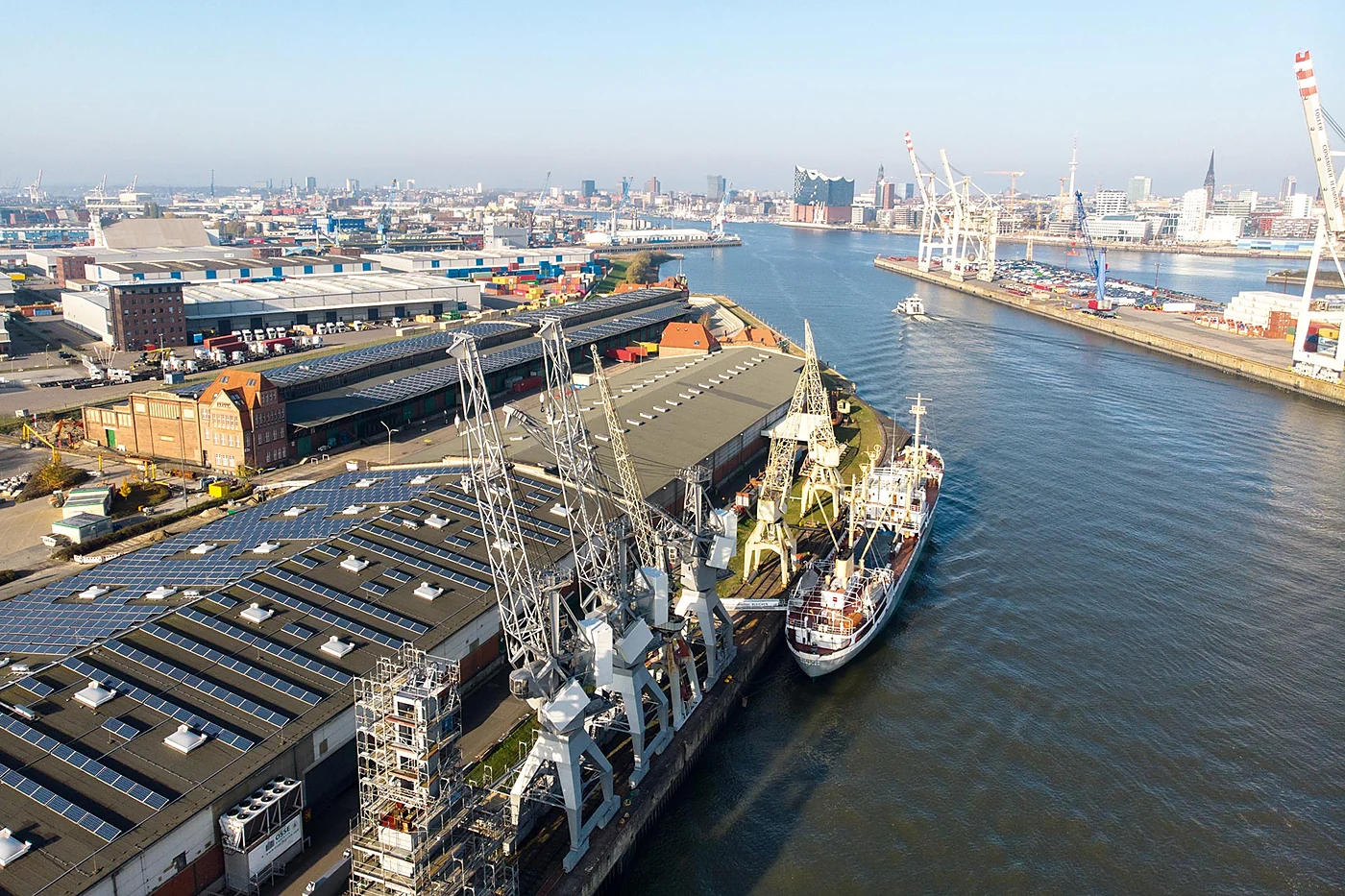 Luftaufnahme der 50er Schuppen auf dem Kleinen Grasbrook mit Kränen, einem Frachtschiff am Kai und der Skyline der Hafen
