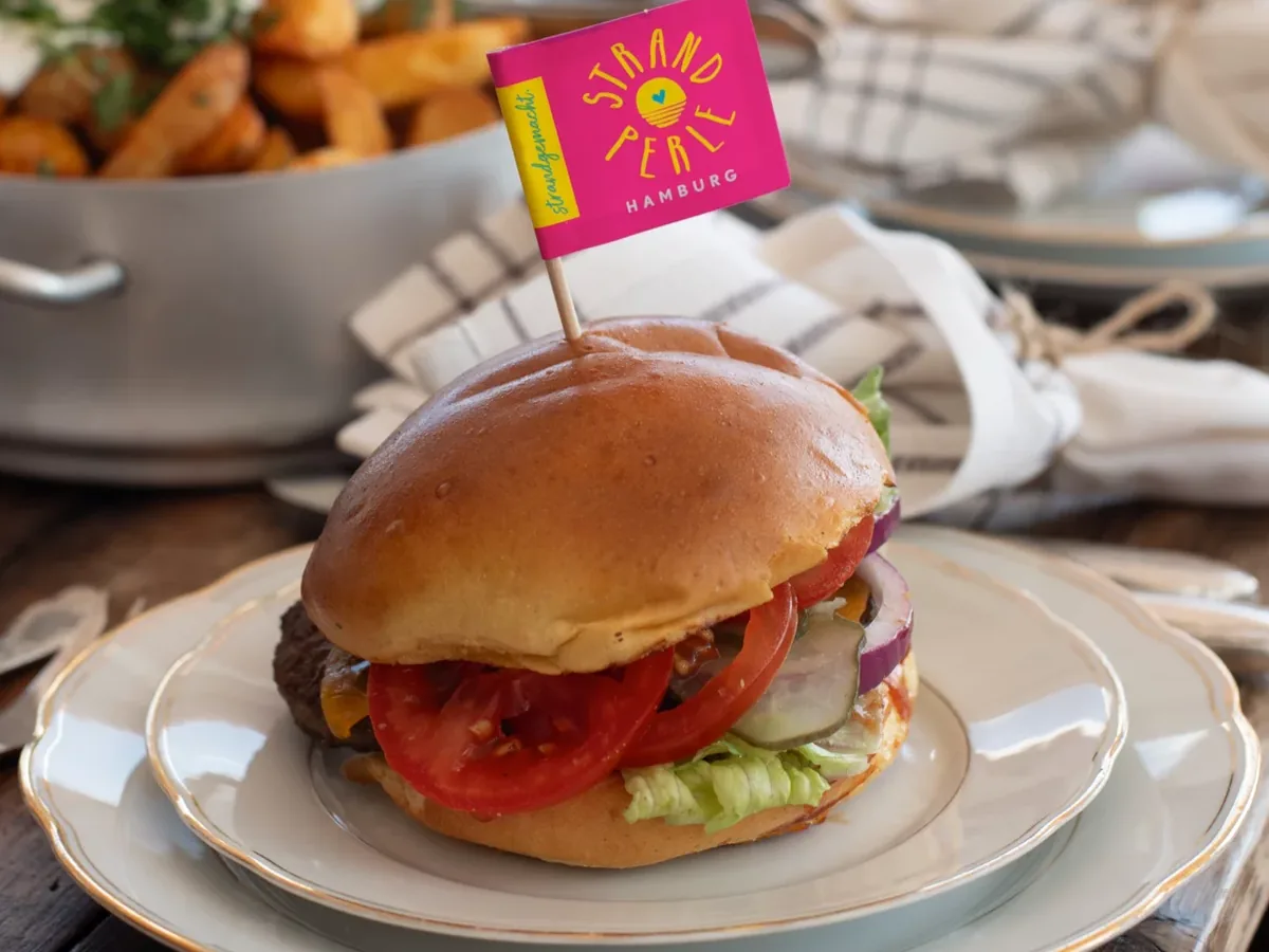 Burger mit Pommes auf einem Teller, dazu ein Fähnchen mit der Strandperlen-Sonne