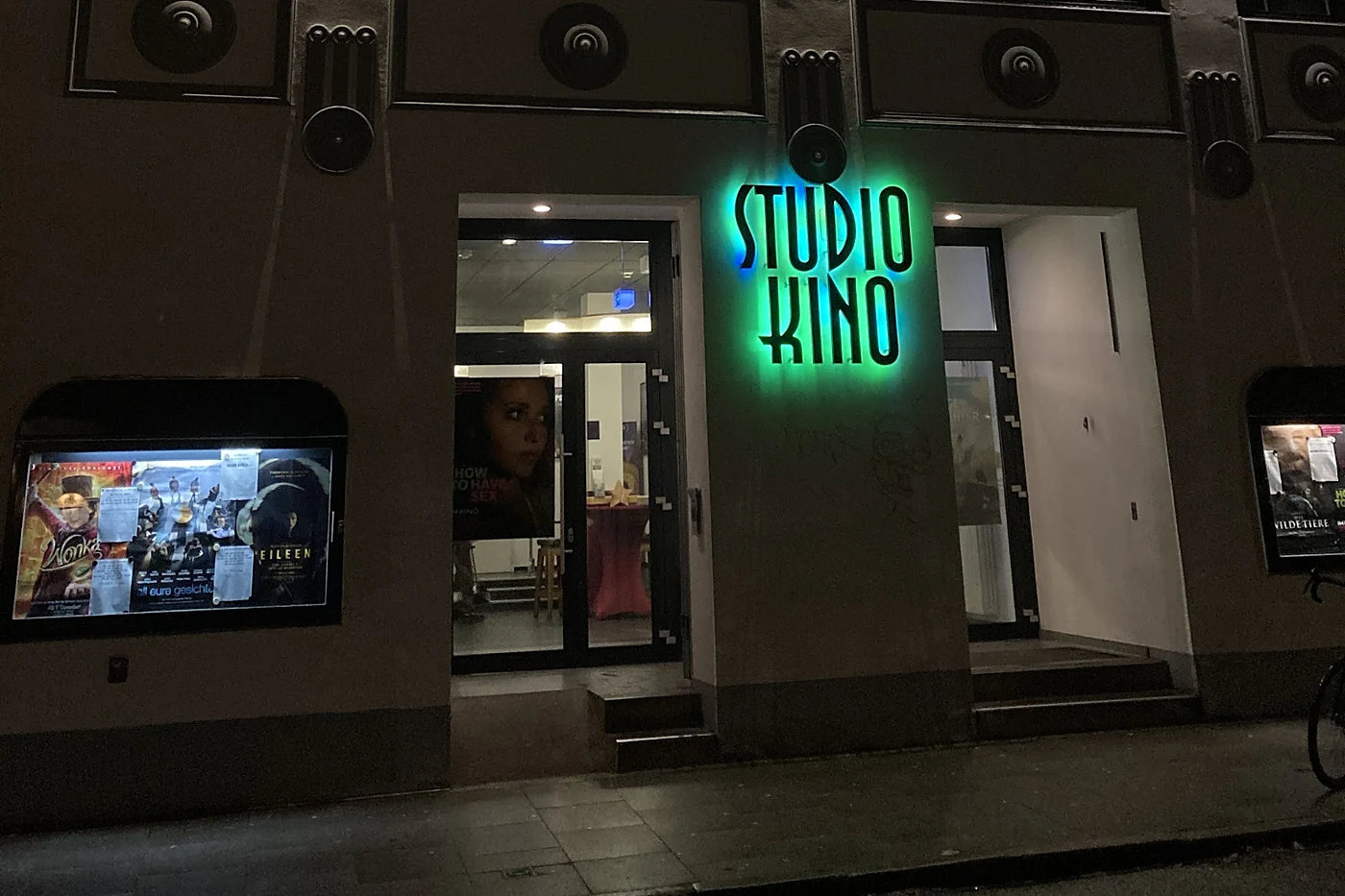 Kinofassade bei Nacht mit grün leuchtendem Schriftzug Studio Kino, offener Eingangstür und Schaukästen