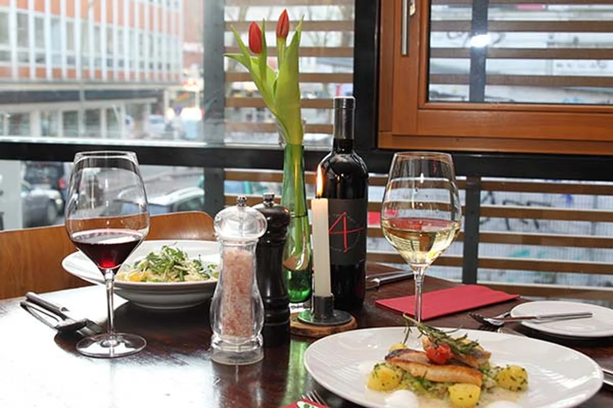 Gedeckter Tisch am Fenster mit einer Schale Pasta, einem Teller gebratenem Fisch sowie Rot- und Weißwein