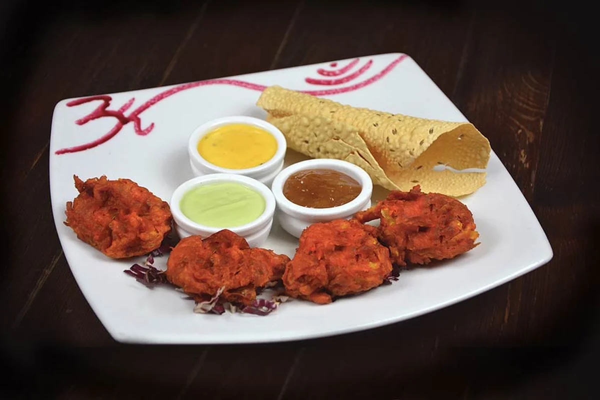 Gemüse-Pakora mit drei Dips und Papadam auf einem weißen Teller mit aufgemaltem Om-Zeichen