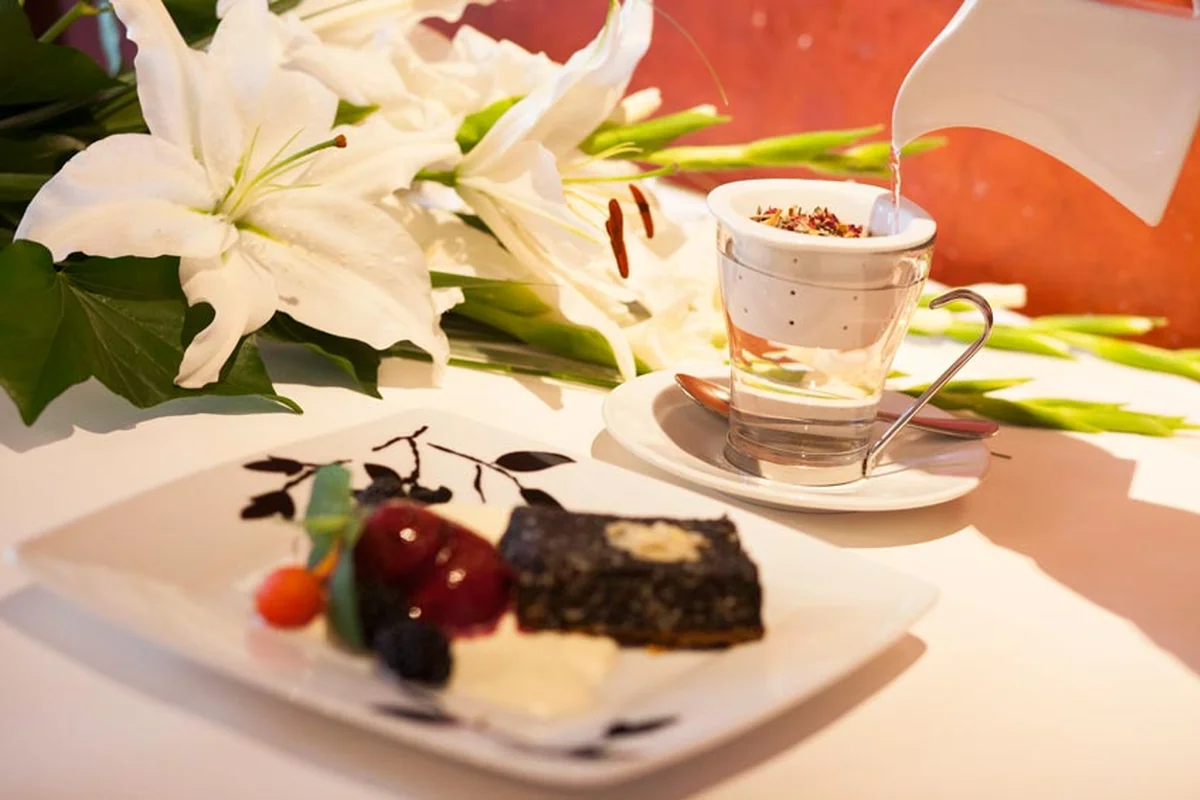 Stück Mohnkuchen mit Beeren, dahinter ein Glas Tee mit frischem Ingwer und weiße Lilien