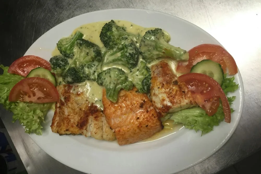Gebratene Fischfilets mit Brokkoli in heller Sauce, Tomate und Gurke auf einem ovalen Teller