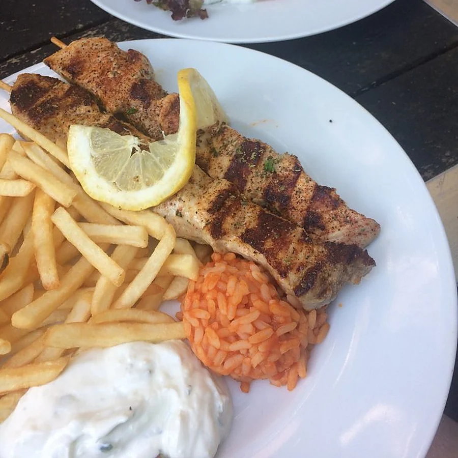 Gegrillter Spieß mit Pommes frites, Tomatenreis, Tzatziki und Zitrone auf weißem Teller