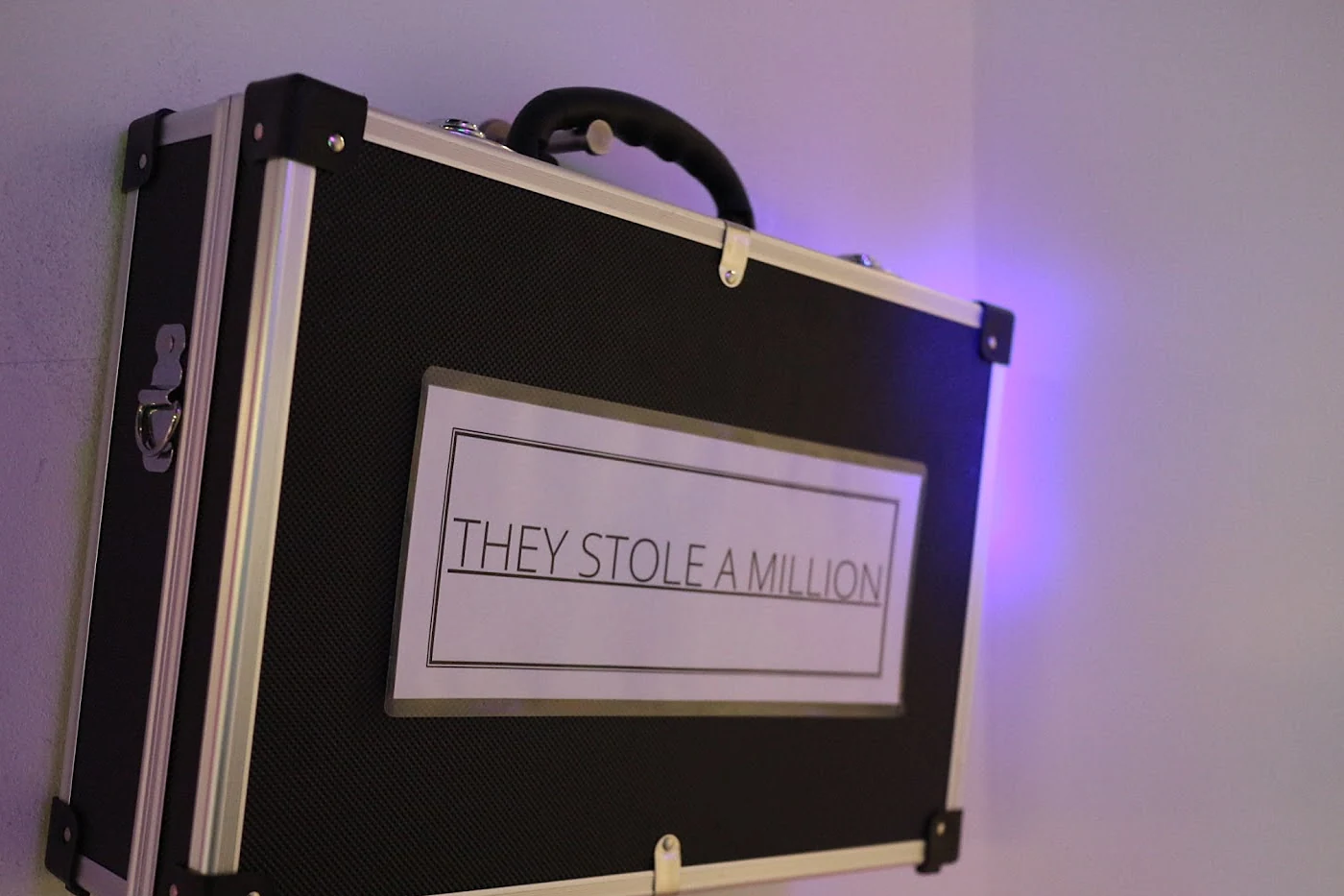 Schwarzer Aluminiumkoffer an einer violett beleuchteten Wand mit dem Schriftzug They stole a million
