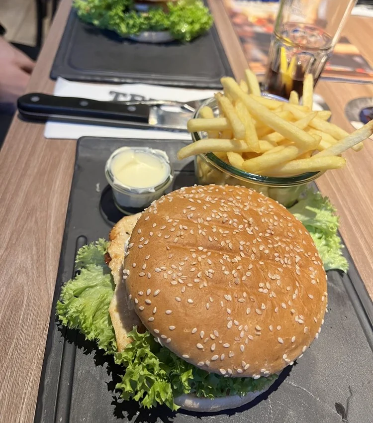 Burger im Sesambrötchen mit Salat auf schwarzer Platte, dahinter Pommes frites im Metalleimer und Dip