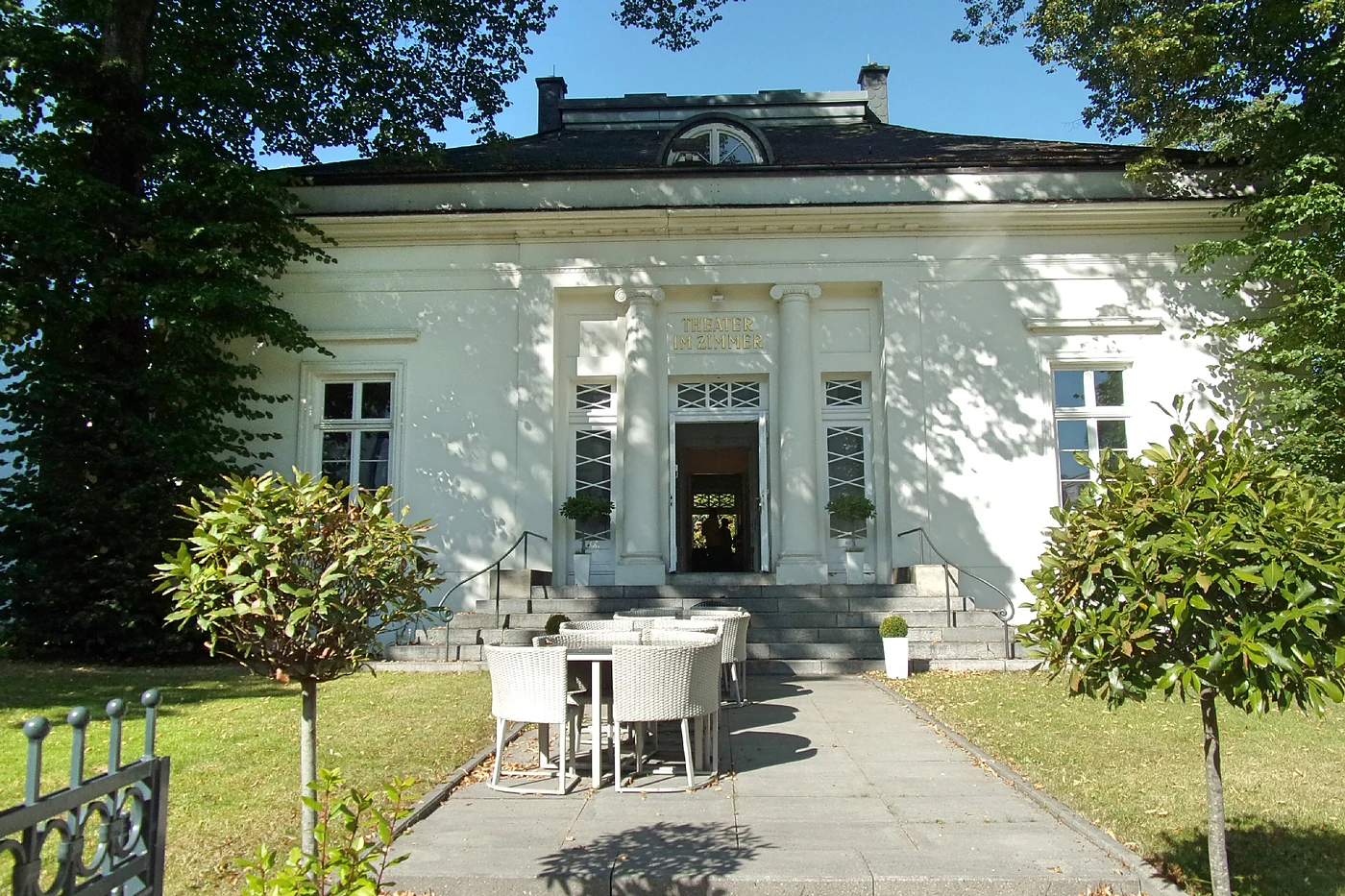 Klassizistische weiße Villa mit Säulenportal, davor Rasen und Terrassentische