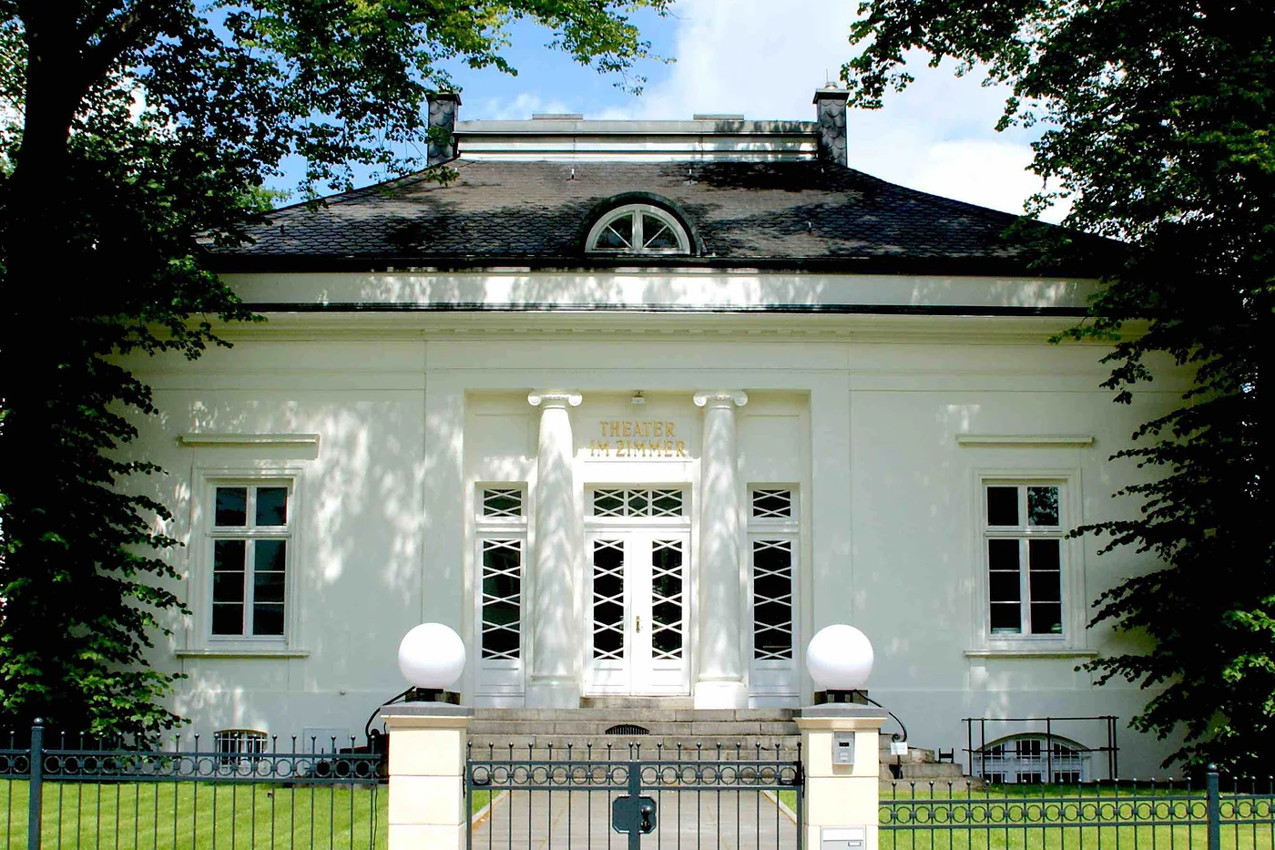 Eingang der Villa hinter schmiedeeisernem Zaun mit zwei Kugelleuchten
