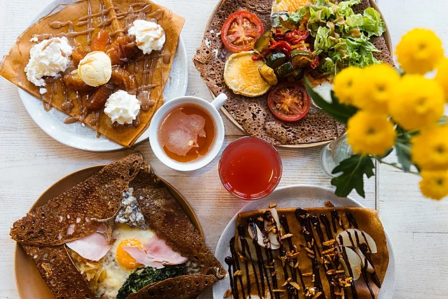 Galettes mit Ei, Schinken und Spinat, mit Salat und Tomaten sowie eine süße Crêpe mit Sahne und Karamell auf weißem Tisch