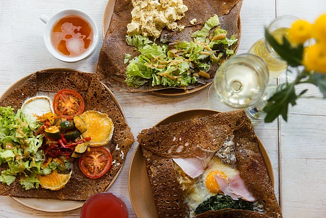 Galettes mit Ei, Schinken, Ziegenkäse und Salat, dazu ein Glas Weißwein und eine Tasse Cidre auf hellem Holztisch