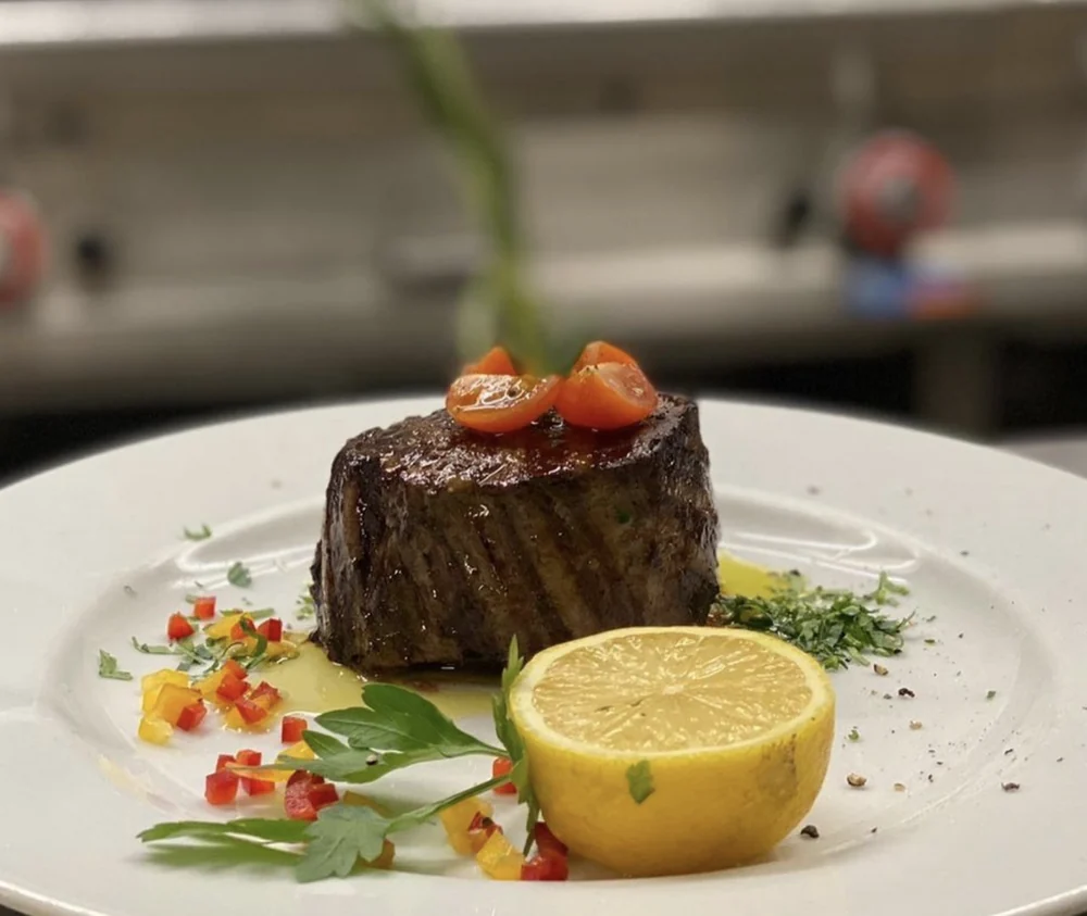 Gegrilltes Rinderfilet mit Kirschtomate, halber Zitrone, Kräutern und Olivenöl auf weißem Teller