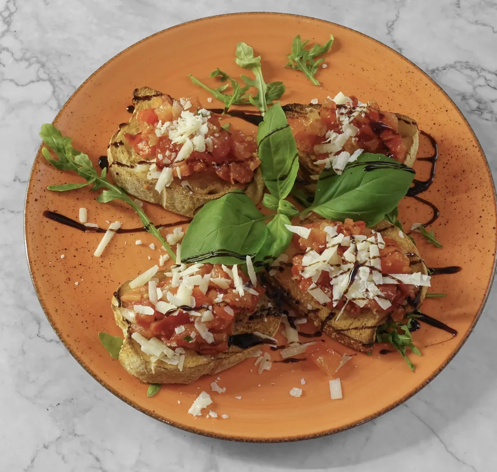 Bruschetta mit Tomatenwürfeln, Parmesanspänen, Basilikum und Balsamico auf orangefarbenem Teller