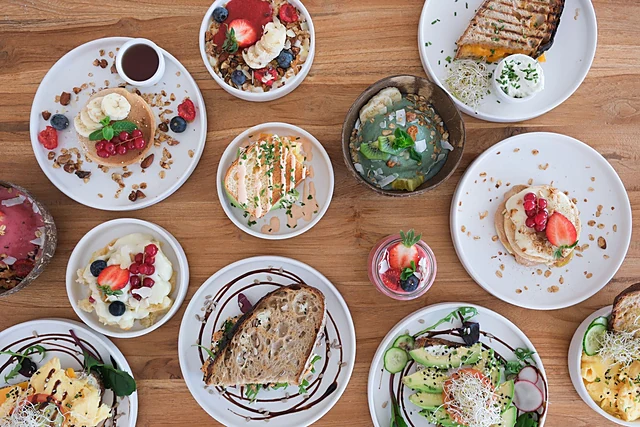 Viele kleine Teller von oben auf Holztisch: Pancakes mit Beeren, Granola-Bowl, Avocado-Brote, Sandwich und Salat