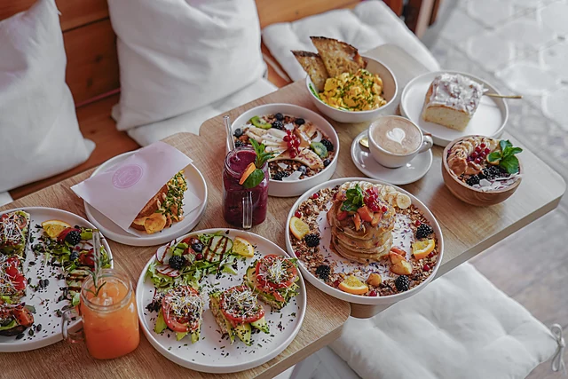 Gedeckter Frühstückstisch mit Avocado-Broten, Pancakes mit Früchten, Bowls, Cappuccino, Cinnamon Roll und Limonade