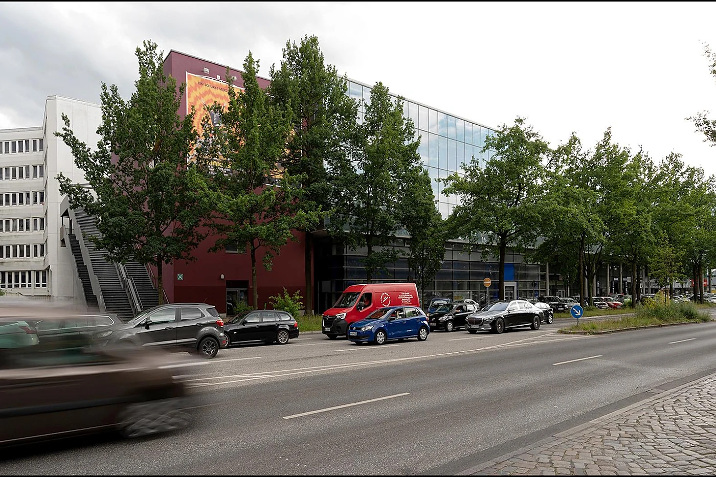 Langgestrecktes Kinogebäude mit durchgehender Glasfassade und rotem Baukörper hinter Straßenbäumen