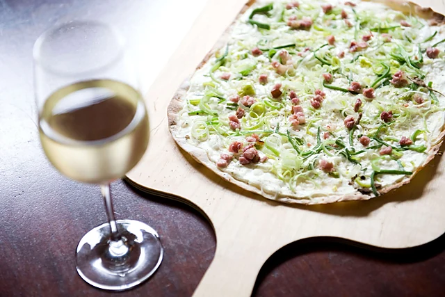 Flammkuchen mit Speck, Lauch und Schmand auf einem Holzbrett, daneben ein Glas Weißwein