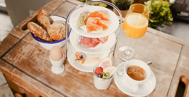 Frühstücksetagere mit Lachs, Brot, Obst und Käse auf Holztisch, dazu gekochtes Ei, Tee und Orangensaft