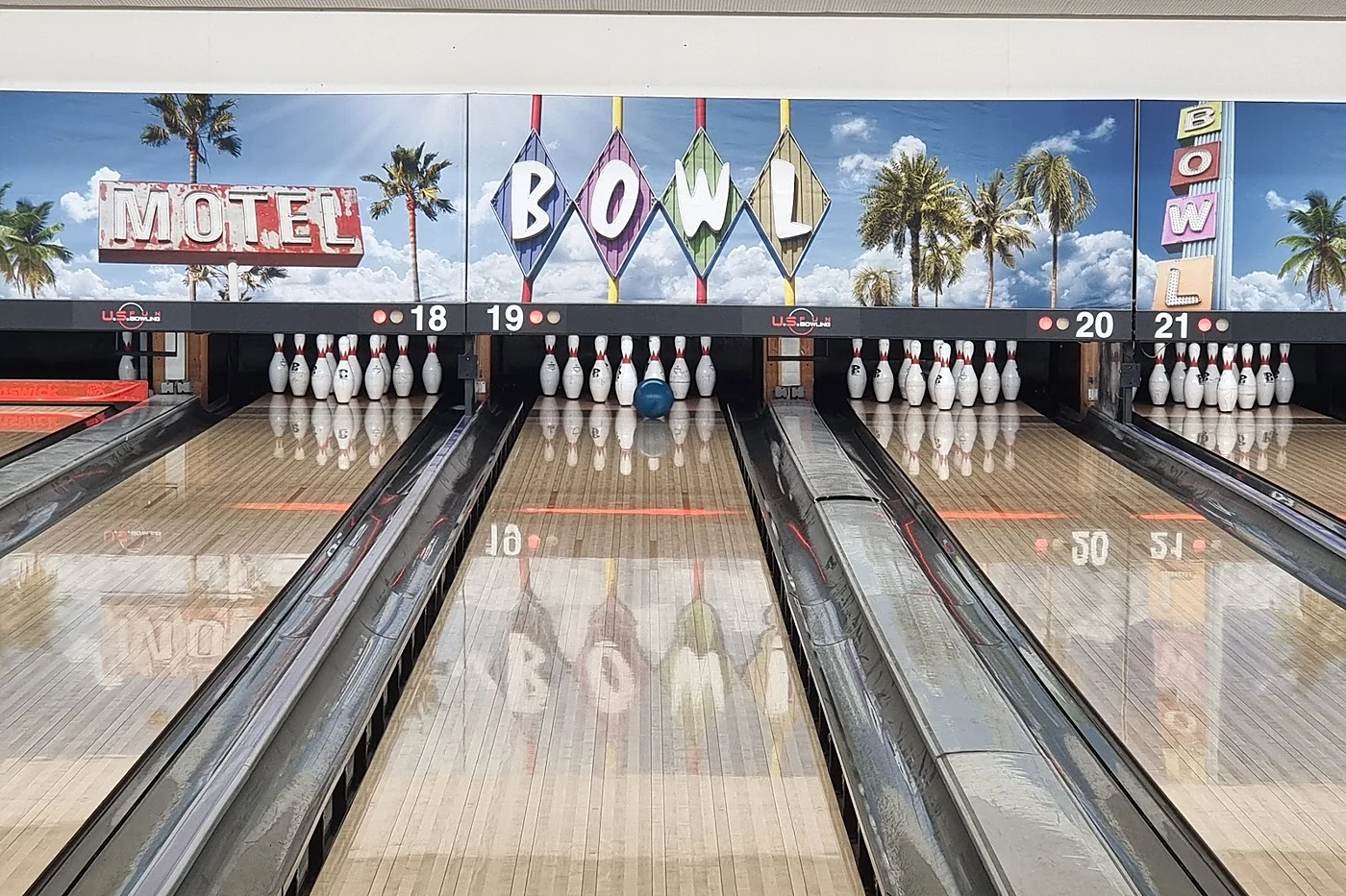 Vier Bowlingbahnen mit aufgestellten Pins vor einer Wand mit Palmen und den Leuchtschriftzügen Motel und Bowl