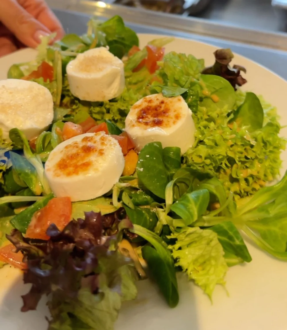 Blattsalat mit vier gratinierten Ziegenkäsetalern und Tomaten auf weißem Teller vor der Antipasti-Theke