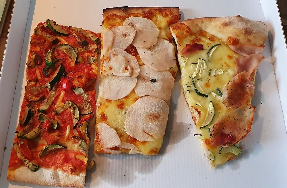 Drei Stücke Blechpizza mit Gemüse, Kartoffeln und Zucchini im Pappkarton