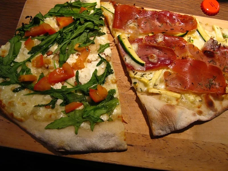 Zwei längliche Pizzastücke auf Holzbrett, eines mit Rucola und Tomaten, eines mit Schinken und Zucchini