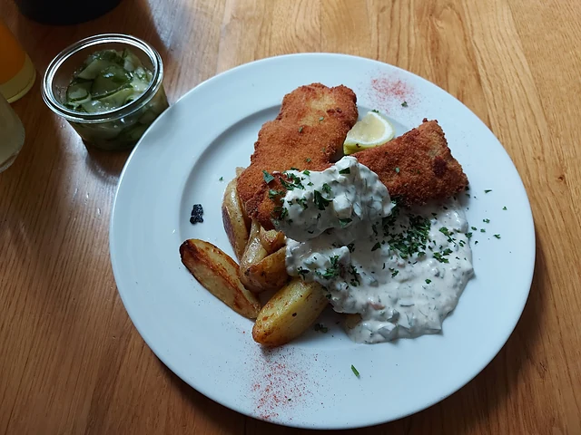 Panierter Backfisch mit Bratkartoffeln und Remoulade auf weißem Teller, daneben ein Glas Gurkensalat