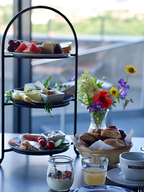 Dreistöckige Etagere mit Obst, Käse, Aufschnitt und Brötchenkorb auf einem Tisch vor der Glasfront, daneben Wiesenblumen, Joghurt im Glas, Saft und eine Waterkant-Tasse