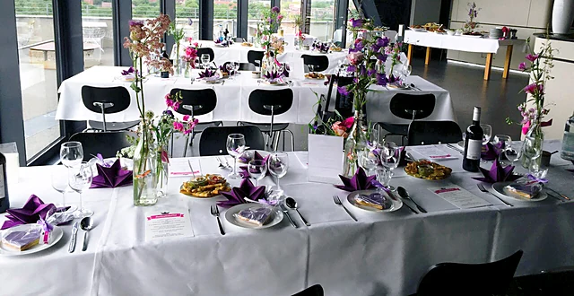 Festlich gedeckte lange Tafel mit weißen Tischdecken, Gläsern, Tellern mit Speisen und lila Blumendekoration vor der Fensterfront mit Blick über Hamburg