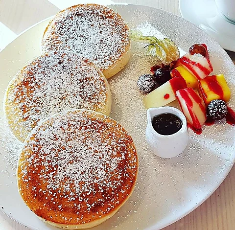 Drei dicke Pancakes mit Puderzucker, daneben Obstspieße mit Beerensoße und ein Kännchen Sirup auf weißem Teller