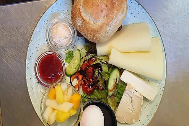Frühstücksteller von oben mit Brötchen, drei Käsesorten, Ei, Salat, Gurke, Obst, Marmelade und Aufstrich