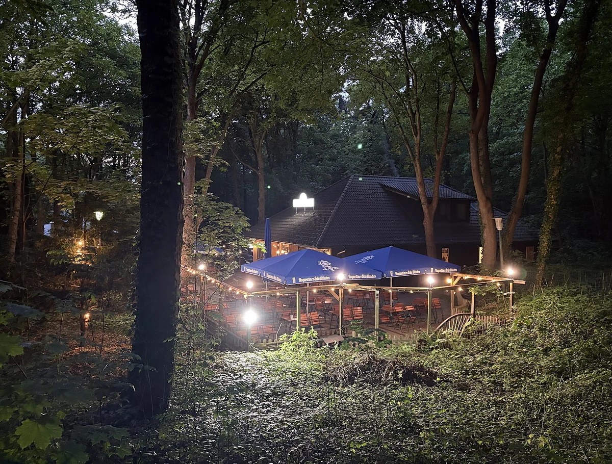 Beleuchteter Gastgarten mit blauen Sonnenschirmen und Lichterketten zwischen hohen Bäumen