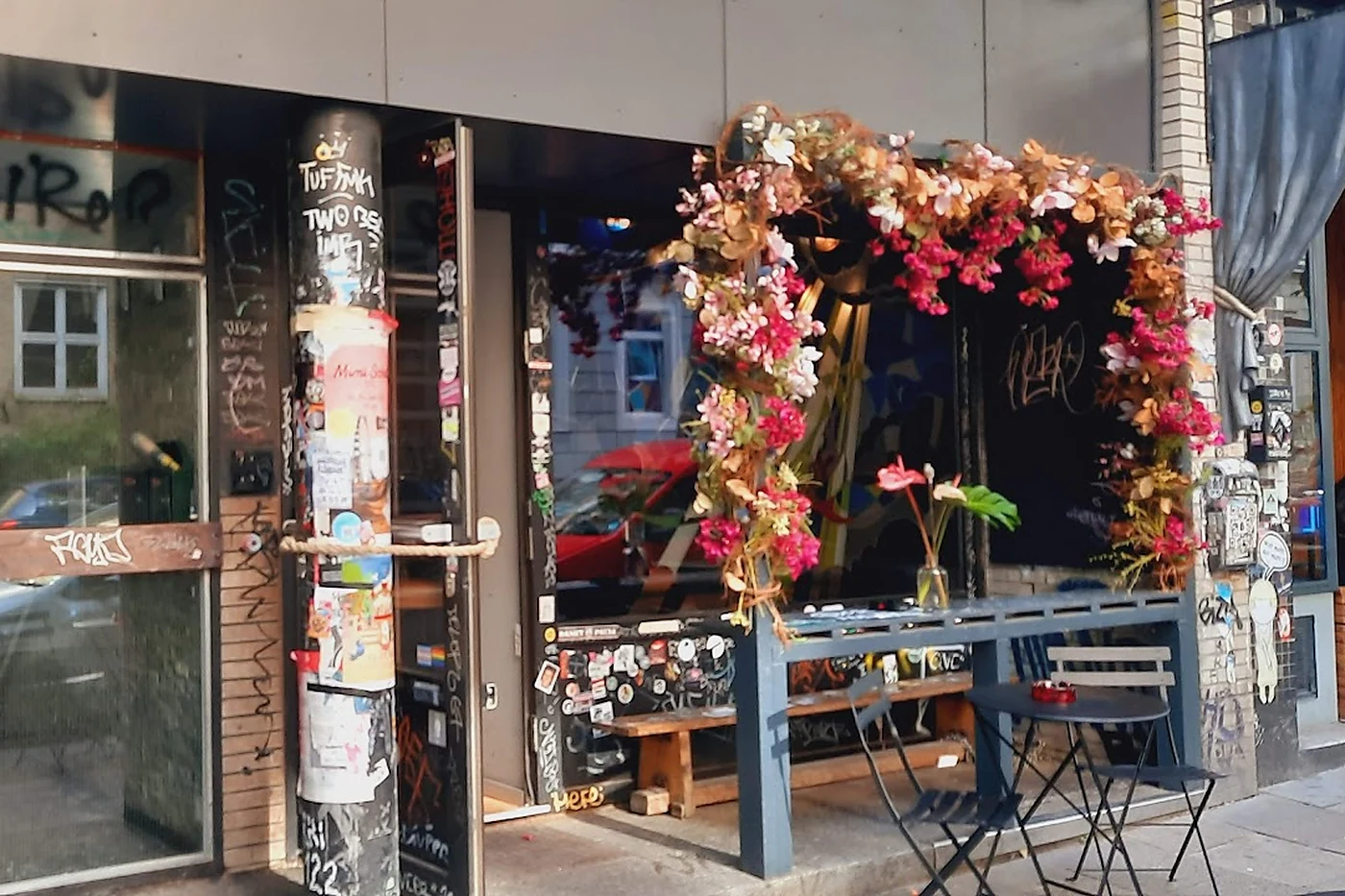Fassade der Walrus Bar in der Wohlwillstraße mit Blumenbogen, beklebter Litfaßsäule und Bistrotischen